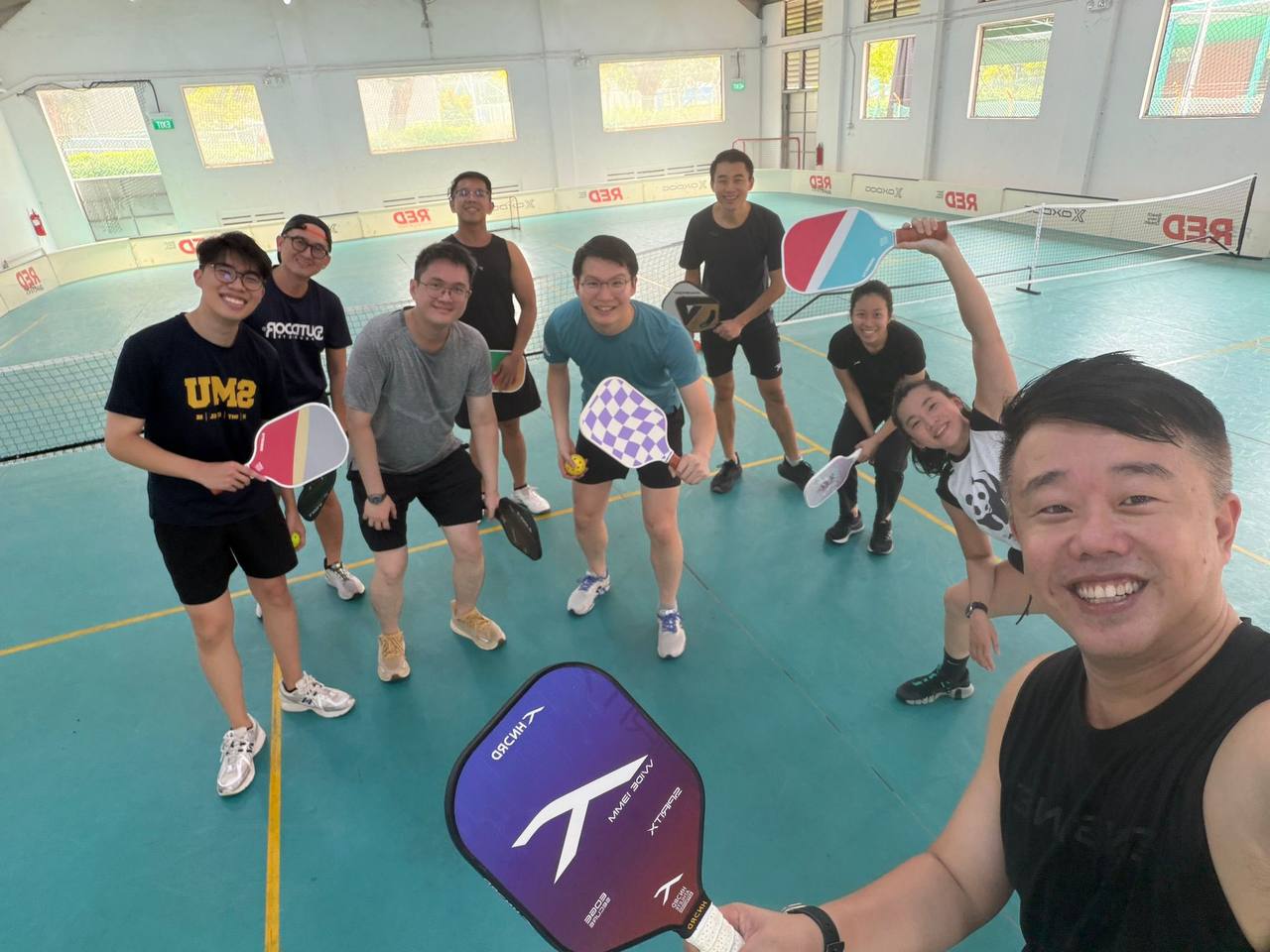 Jordan and a group of friends holding pickleball paddles and posing for a selfie on an indoor court