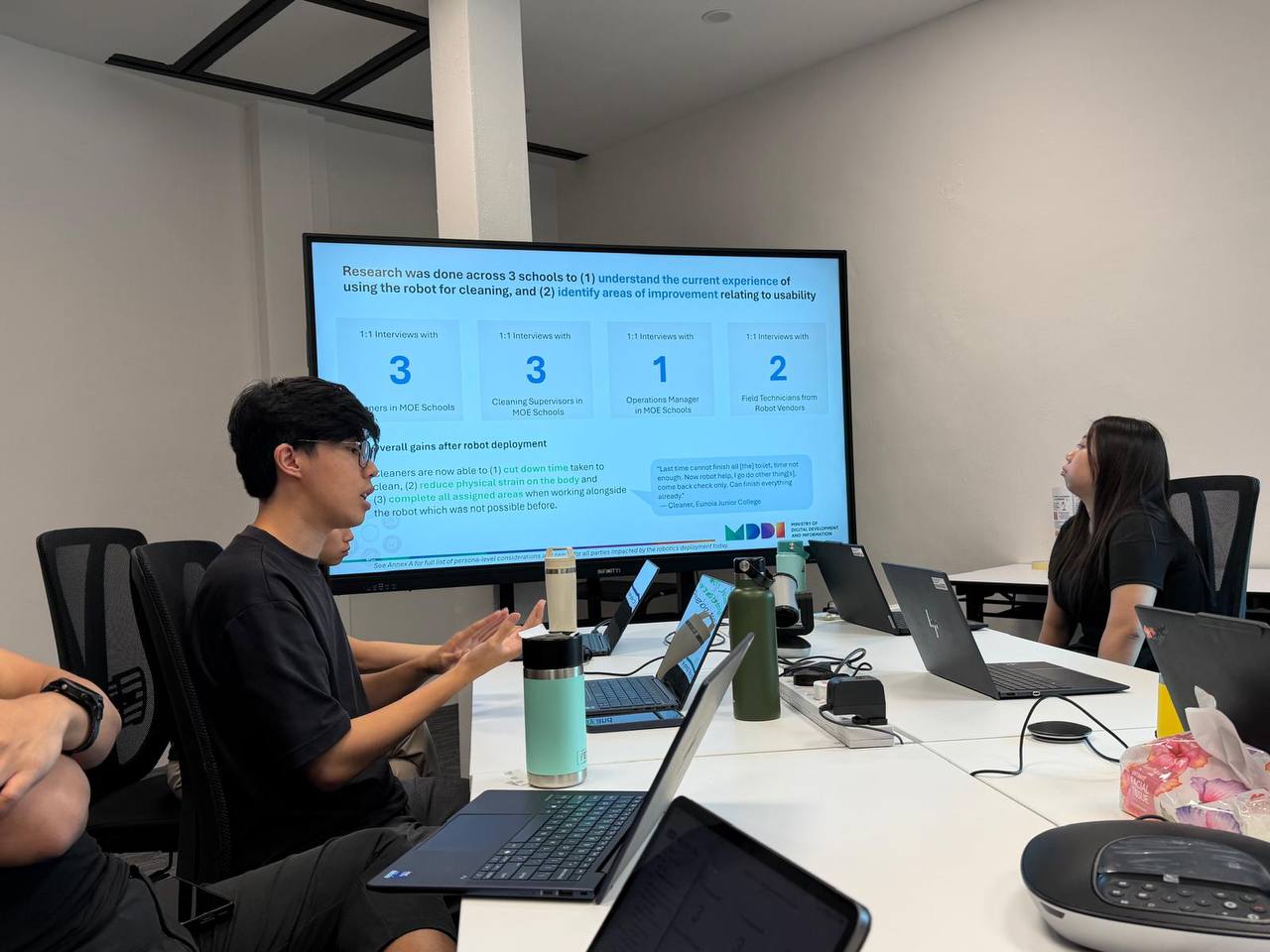 Jordan presenting research findings to a room of government stakeholders, with a summary slide of interview coverage and a cleaner's quote shown on screen